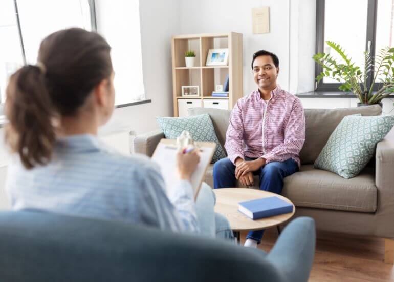 Um homem sentado sorridente em um sofá enquanto conversa com uma profissional, que está de costas para a câmera, em um ambiente claro e acolhedor que parece ser um consultório.