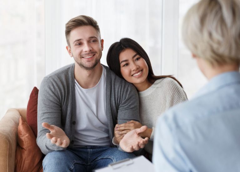 Um casal jovem sorrindo e conversando com uma pessoa cujas costas estão voltadas para a câmera, possivelmente em uma sessão de aconselhamento ou terapia de casal.