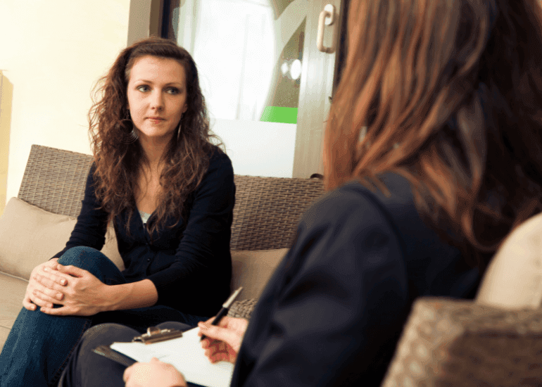 Uma mulher sentada em um sofá conversa com uma profissional que está tomando notas, possível contexto de consulta terapêutica ou coaching.
