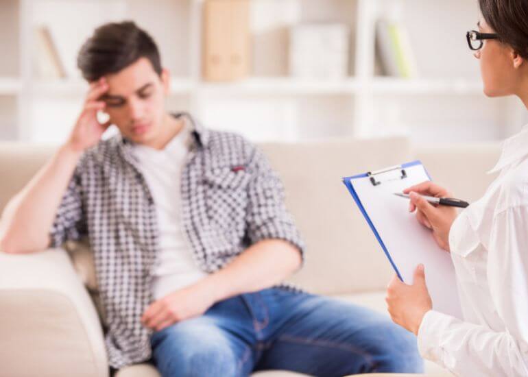  Um homem jovem sentado no sofá com a mão na cabeça parecendo preocupado, enquanto uma profissional de pé ao lado segura um prancheta e caneta, indicando uma sessão de aconselhamento ou entrevista.