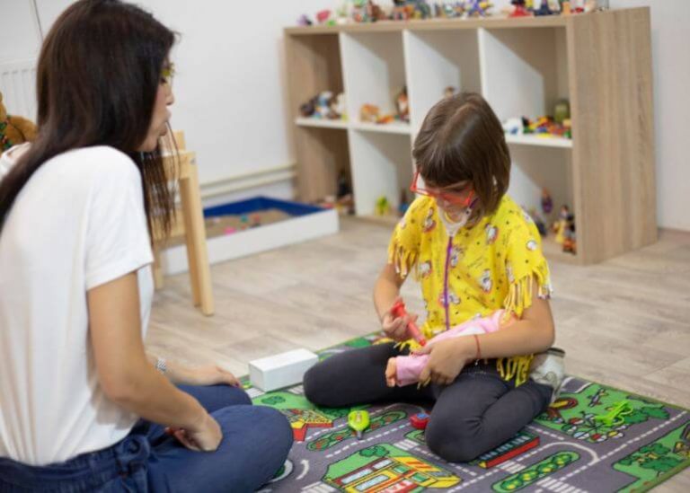 Uma criança brincando com brinquedos no chão de uma sala de jogos enquanto uma mulher adulta observa sentada ao lado.