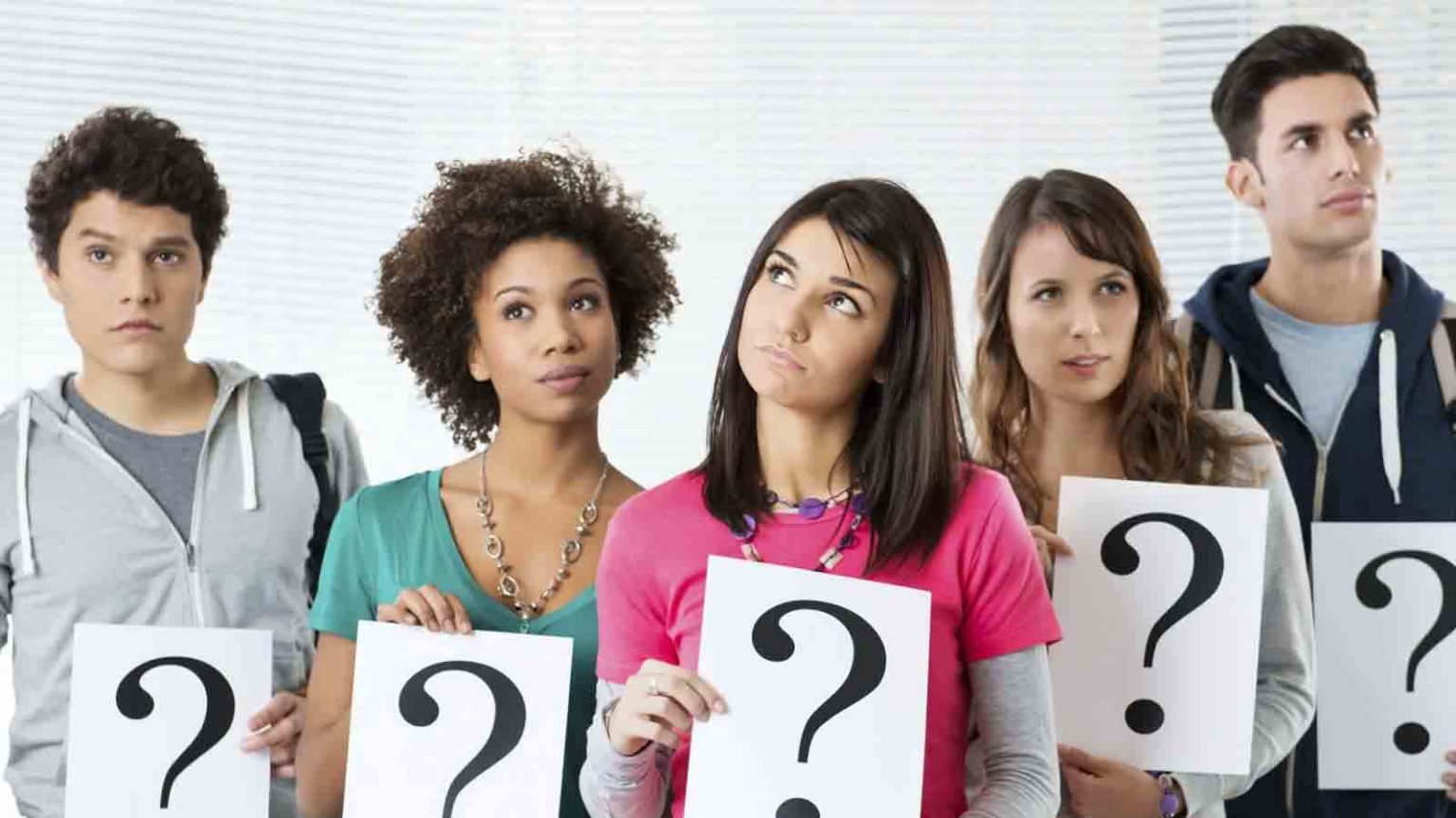 Um grupo de cinco jovens com expressões pensativas segurando cartazes brancos com pontos de interrogação pretos, sugerindo incerteza ou busca de respostas.