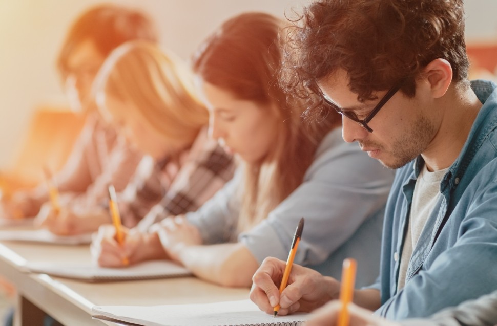 Um grupo de estudantes concentrados escrevendo em cadernos em uma sala de aula, com foco em um homem jovem na frente, simbolizando aprendizado, educação ou realização de uma prova.